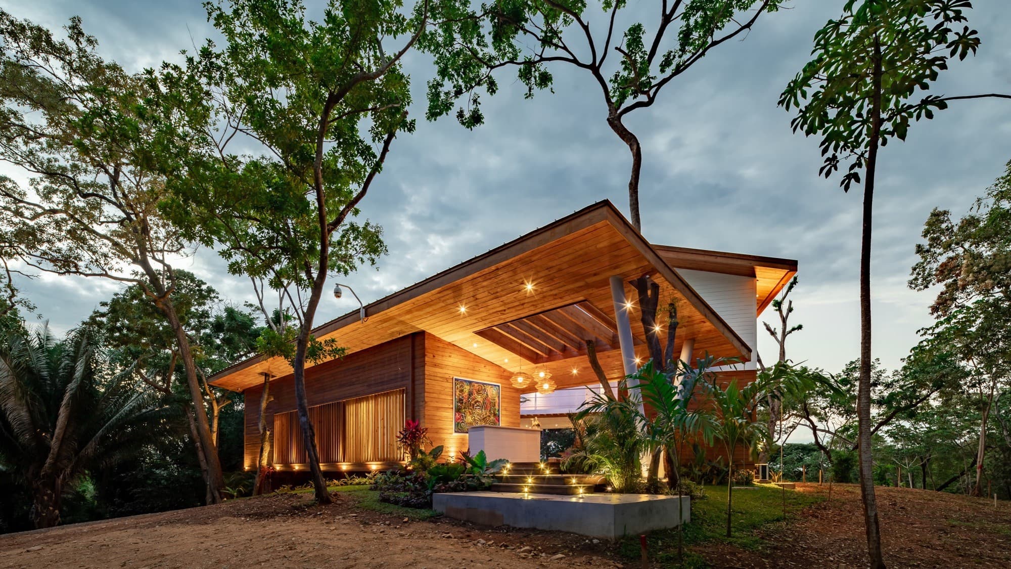 Modern house with a wooden exterior and expansive overhanging roof, beautifully lit at dusk. The home is surrounded by lush tropical plants and trees, creating a serene and natural atmosphere.