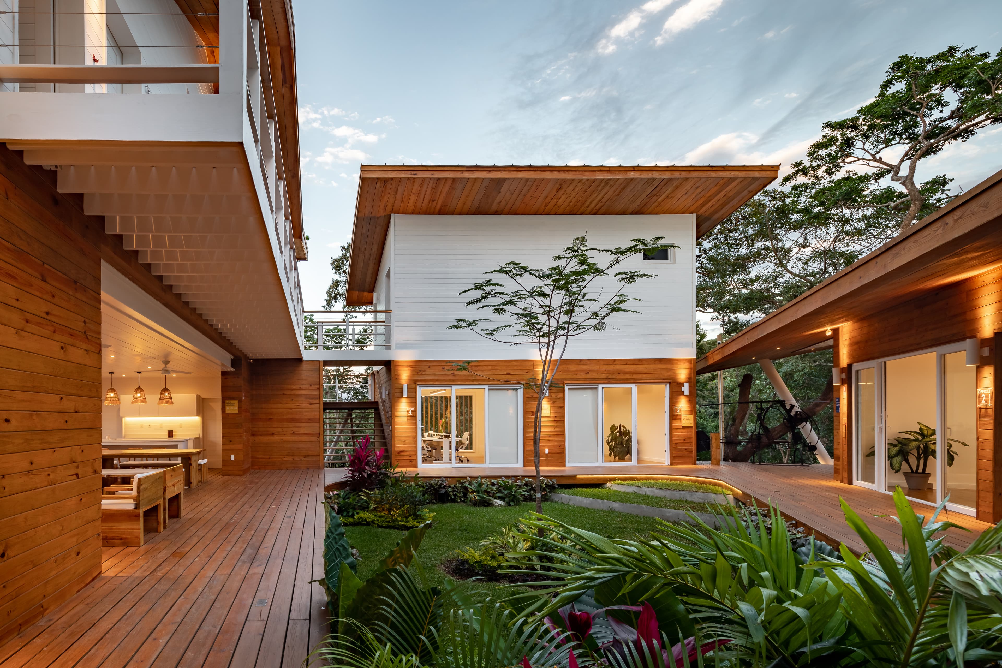 A contemporary house with a wooden exterior featuring an open courtyard with lush plants and trees. The house has large glass doors and windows, allowing natural light to illuminate the indoor spaces, and the wooden deck connects various parts of the home, creating a seamless indoor-outdoor living experience.