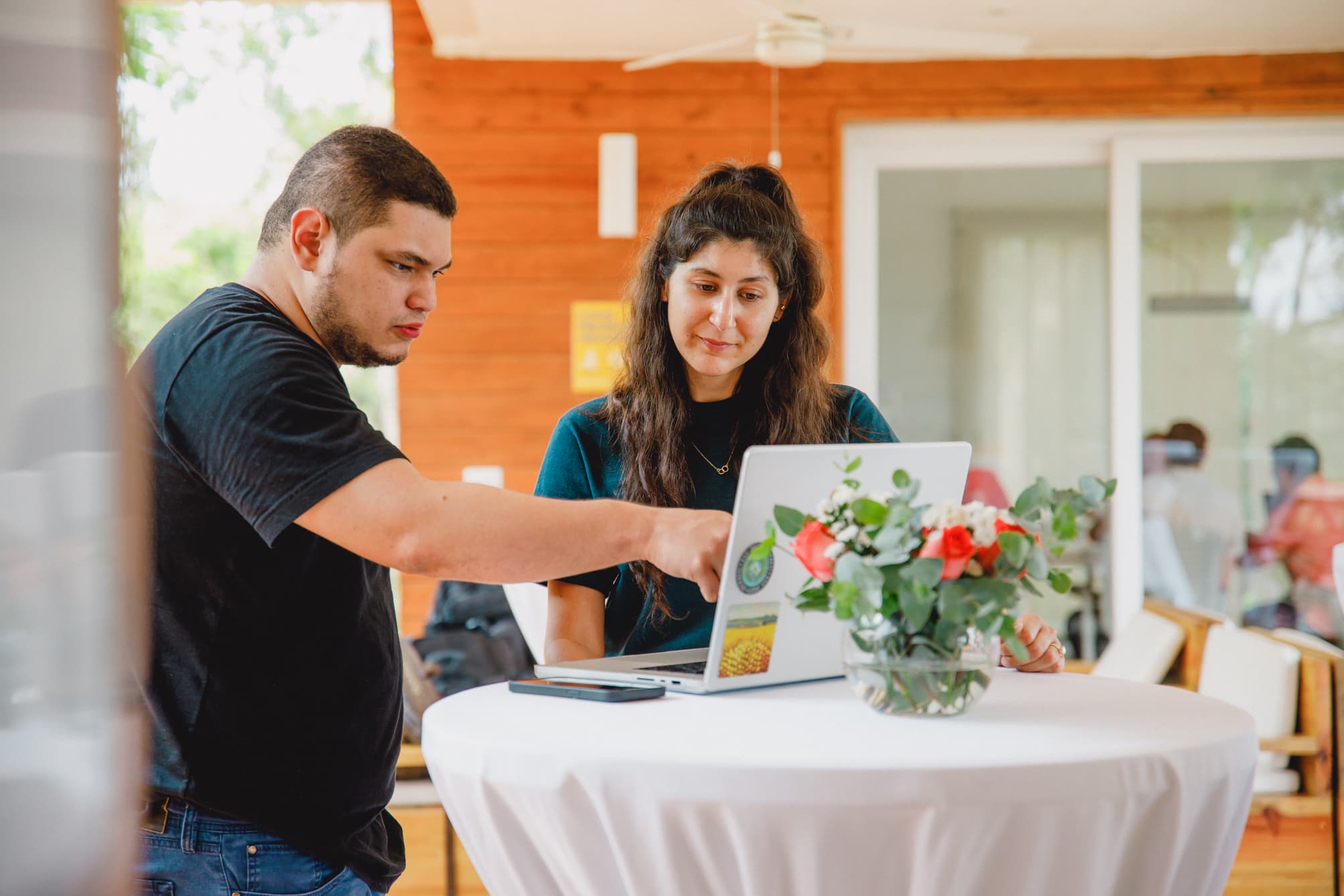 Two people standing at a high table, with a laptop open in front of them. One person is pointing at the screen while the other looks on attentively. A vase with flowers decorates the table, and the background features a wooden interior, creating a casual and collaborative setting.