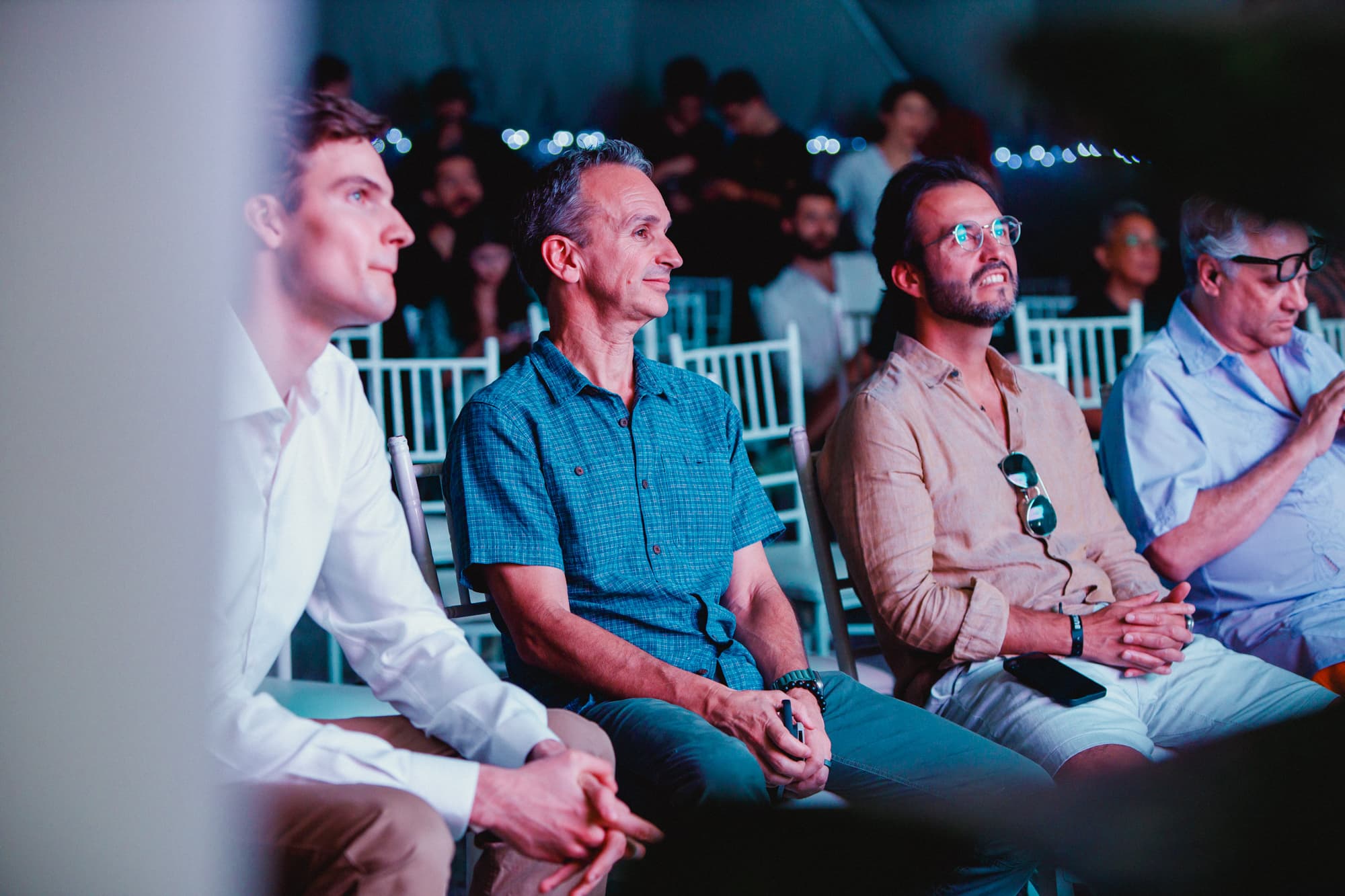 Three men seated in a row, attentively watching an event, with a crowd in the background. The setting appears to be indoors with a focus on the front row attendees.